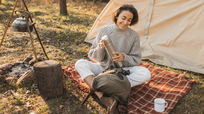 Woman sits on camping blanket while holding thermos