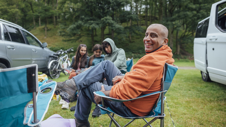 Smiling man and three kids sit in camping chairs in the woods.
