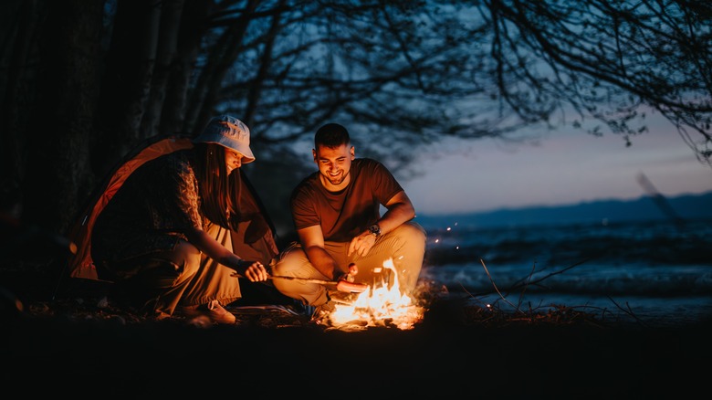 Two people crouch near a campfire in the dark