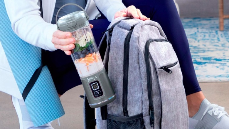 A low-angle image of an unidentified woman in workout clothing, carrying a yoga mat and placing her Ninja Blast Portable Blender inside her backpack.