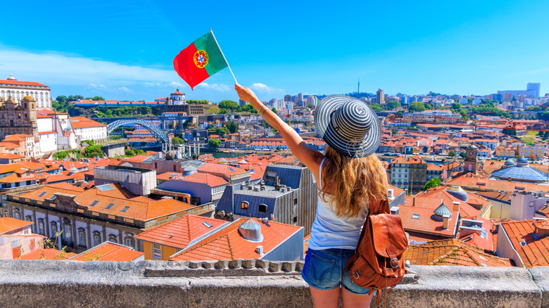 Woman in blue and white hat and backpack waves Portuguese flag over the red rooftops of a city