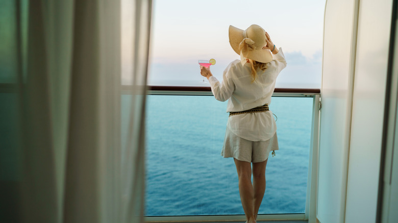 A traveler stands on the balcony of a cruise ship, overlooking the water with a drink in hand