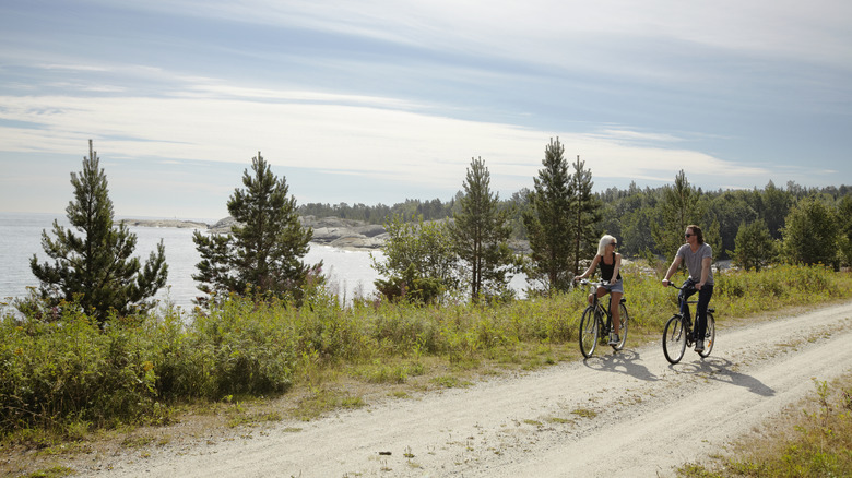 Man and woman cycling on a trail by a tree-lined lake on an overcast day.