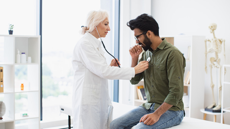A female doctor gives a male patient a respiratory exam, holding her stethoscope to his chest while he coughs