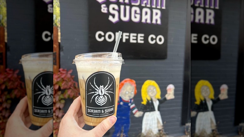 A hand holds up an iced coffee drink in front of a horror-themed mural