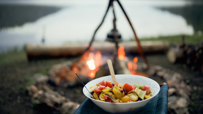Fruit salad in a white bowl in the foreground with a faded background of a campfire
