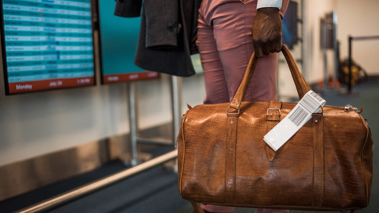 A man stands at an airport with a jacket and a carry-on bag in his hand at an unidentified airport