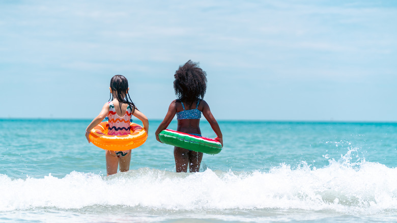 Two happy children with their backs to the camera hold donut floaties as they prepare to run out into the open ocean in front of them.