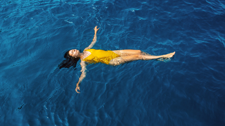 A young woman in a yellow one-piece swimsuit floats peacefully on her back in the open ocean.