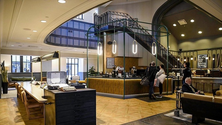 Lobby of Joinery Hotel, with wood floors around a check-in desk in front of a black-railed staircase in a high-ceilinged room
