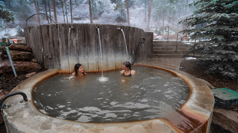 Two people enjoying a soak in a double-tier hot springs with water cascading and snowy trees in the background at Durango Hot Springs Resort & Spa