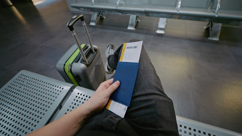 A passenger waits by a gate with a suitcase and boarding pass