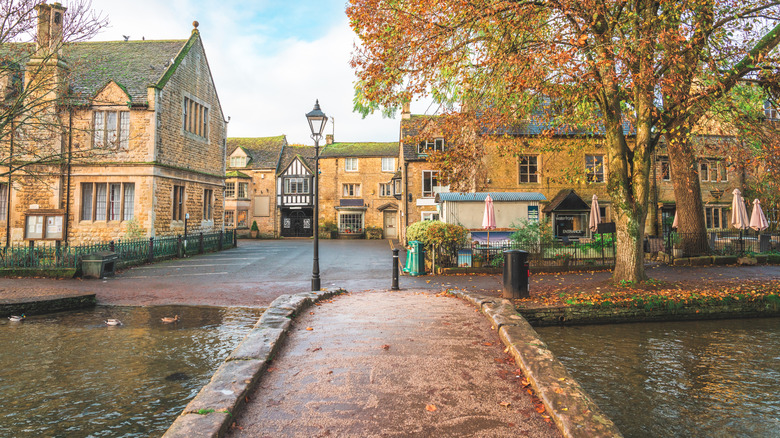 A pedestrian bridge over a river leads to honey-colored cottages on a fall day in Bourton-on-the-Water