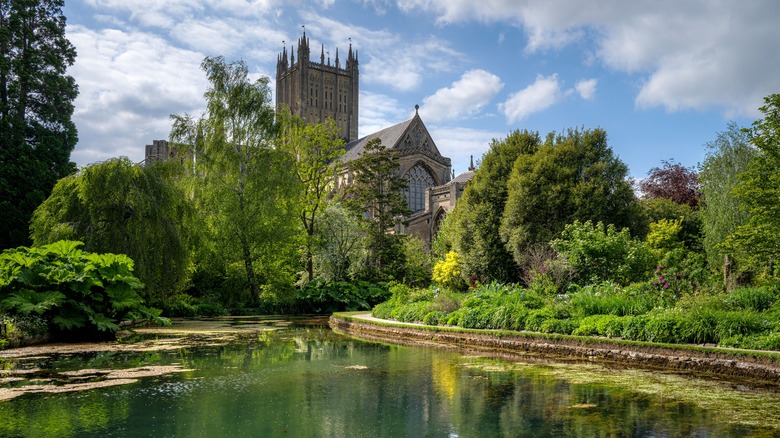 Wells Cathedral surrounded by lush greenery and a tranquil pond under a partly cloudy sky in Somerset, England