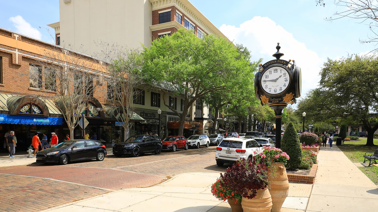 Street view of Park Avenue in Winter Park, Florida with cars parked along the streets, a clock at the front and storefronts behind the cars on a brick-paved street.