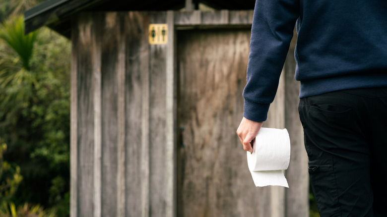 A man holding a roll of toilet paper with an outhouse in the background.