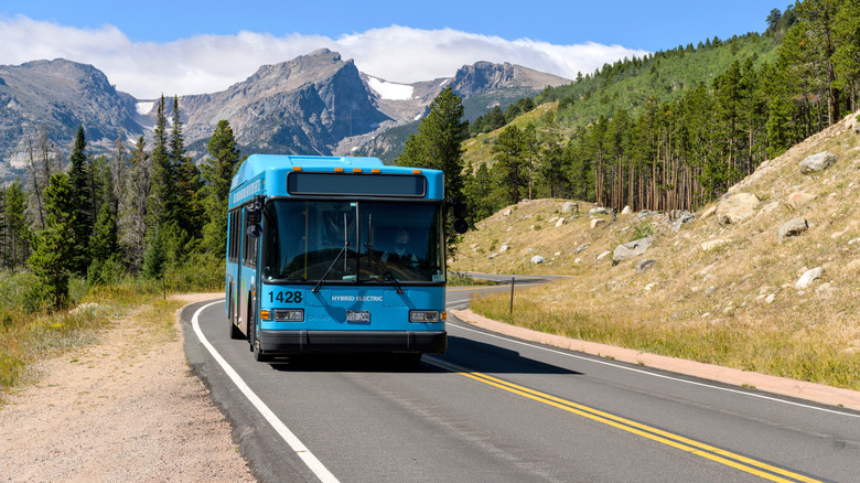 a blue bus on a winding road with forest and mountains in background