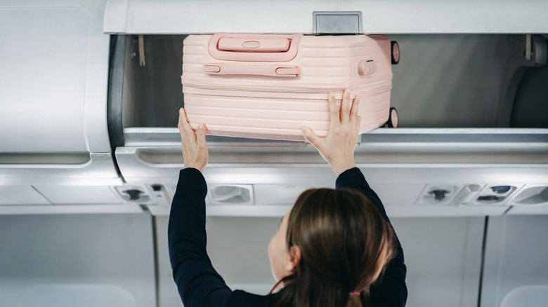 A woman storing her pink luggage in an overhead bin