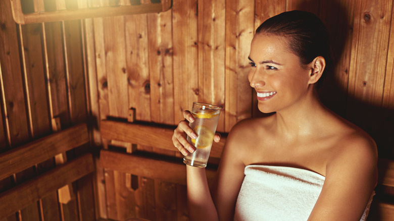 A smiling woman drinking a glass of lemon water while in a sauna wrapped in a towel