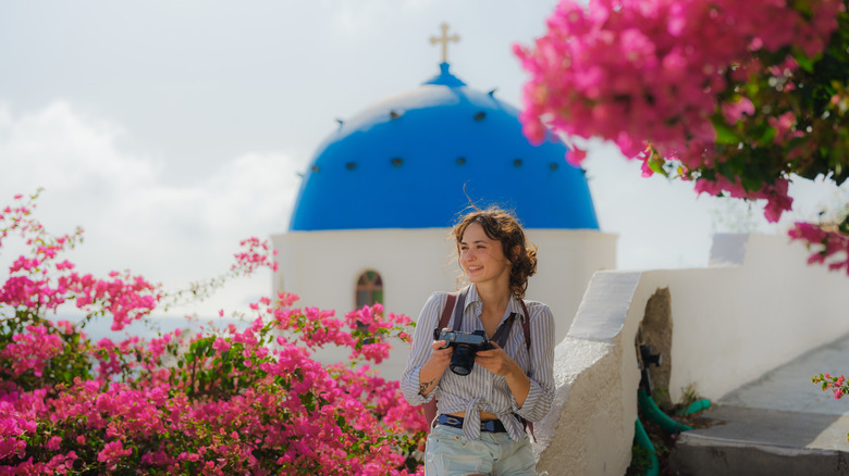 A woman holding a camera with a blue-domed church in the background, surrounded by bougainvillea in Greece