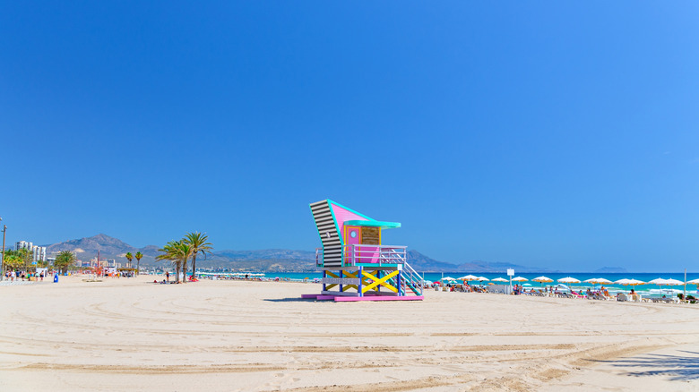 Blue skies over a white-sand beach, with a colorful lifeguard station, loungers, and palm trees