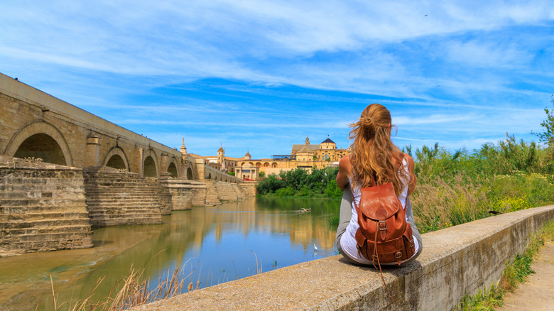 Female solo traveler sitting by a river and old European town in the background