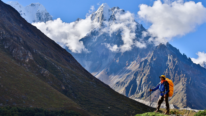 A lone climber with large orange backpack and hat stands with a walking stick before the Kanchenjunga mountain