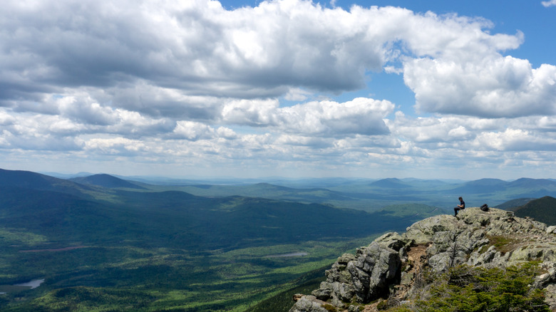 A hiker sits atop a rock outcropping at the summit of Bigelow Mountain Maine with lush forest and mountains cascading in the background.