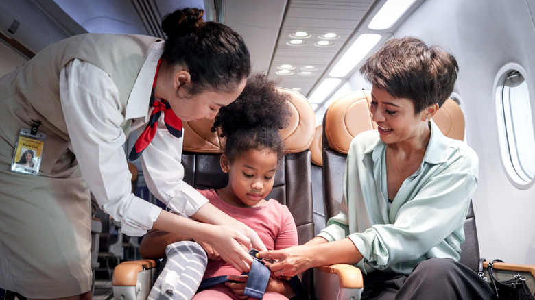 A flight attendant helps a child and her mother fasten her seatbelt