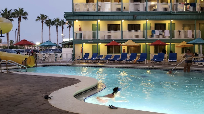 View of people swimming at Sandpiper Beacon Beach Resort pool area at dusk with waterslide on lefthand side and bright yellow guest room building in background