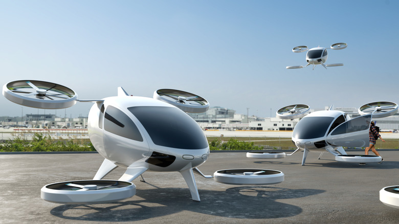Fleet of white electric-powered Vertical Take Off and Landing eVTOL aircraft parked with airport buildings visible in the background
