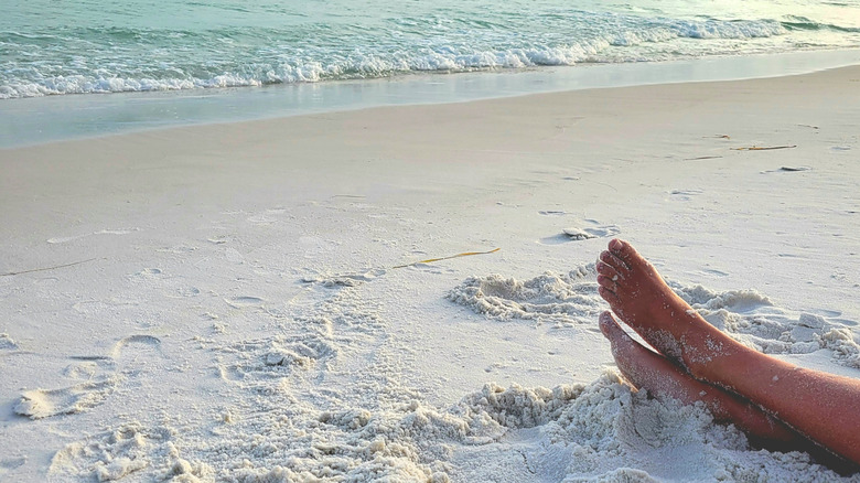 Woman's bare feet on a white sand beach, ocean in the background.