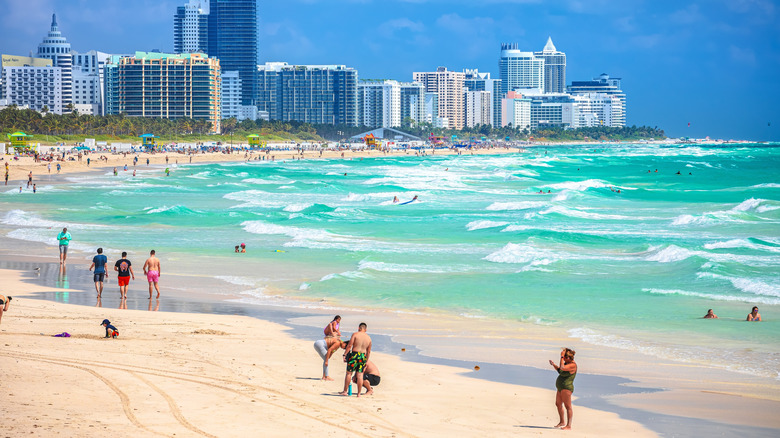 A beach in florida with blue-green water and gentle waves filled with people with big buildings in the background