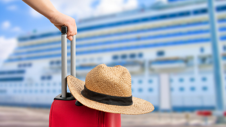 Hand on suitcase with straw hat in foreground and cruise ship in background