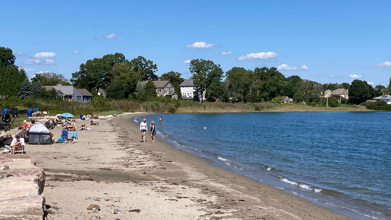 People sunbathing and paddling on North Kingstown Town Beach, with trees and houses in the background