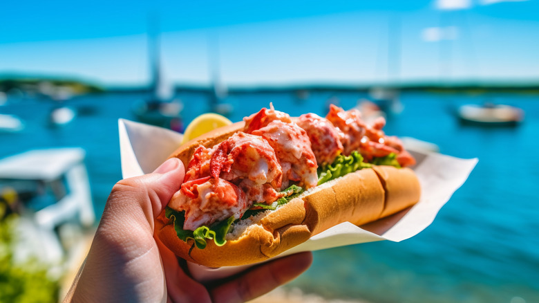 A hand holds up a lobster roll in front of sailboats on a bright day
