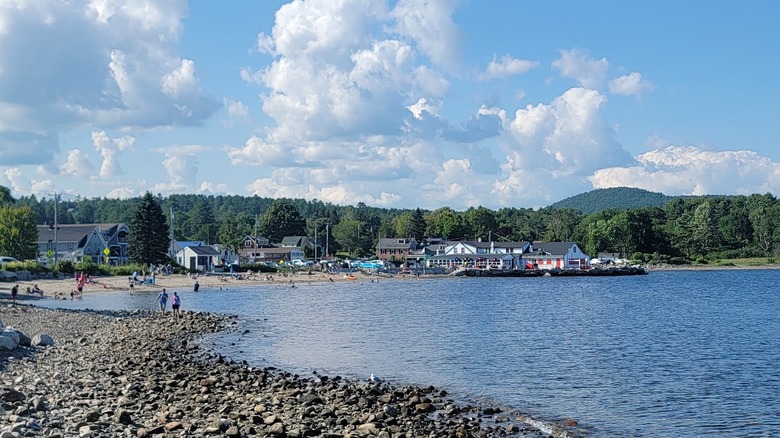 Lincolnville Beach, with a rocky shore in the foreground and people on a sandy beach with buildings behind it further up the shore, surrounded by trees