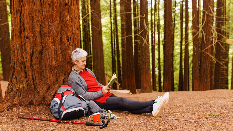 A gray-haired woman with a backpack sitting under a redwood tree reading a book