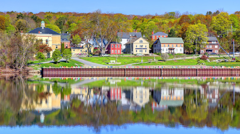 Hallowell viewed from the Kennebec River, with red and yellow brick homes behind green grass and a brown structure along the river bank lined in colorful lawn chairs, with trees in the background under a blue sky