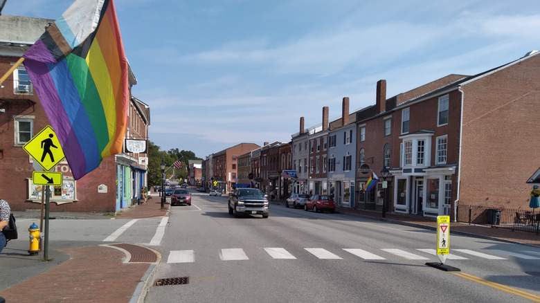 Downtown Hallowell, with cars driving on a street lined in brick sidewalks and storefronts and a rainbow flag in the foreground, under a blue sky