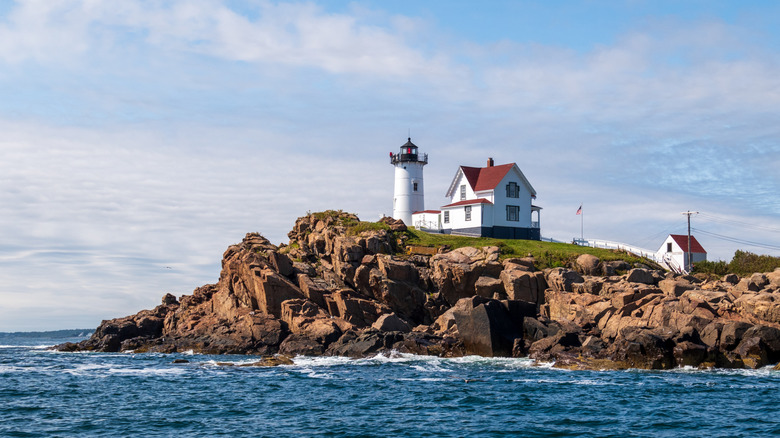 A dark blue shore and a rocky coastline with green carries upon it the white Cape Neddick Nubble Lighthouse and red-roofed dwelling in York County, Maine