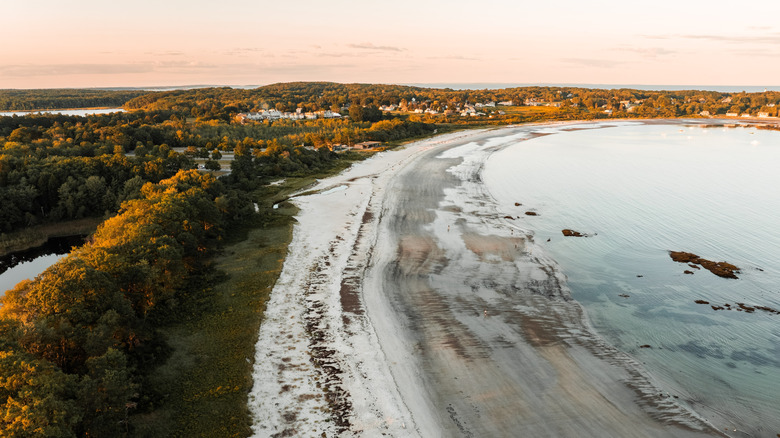 The white sands of Crescent Beach stretch into the waters of Casco Bay in front of evening-lit trees and rolling hills with a peach colored sky