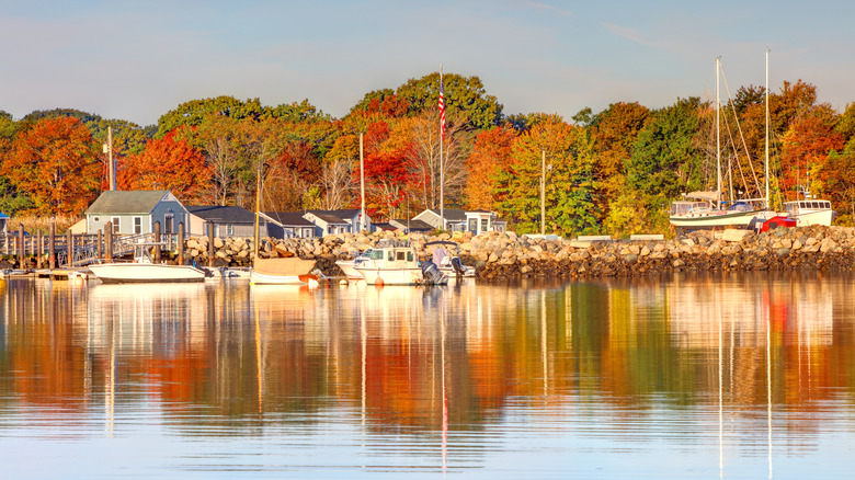 Homes and boats along the harbor in Rye, New Hampshire, with autumn trees in the background reflected in the ocean water in the foreground, under a blue sky