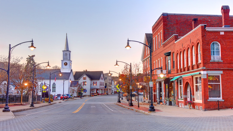 Downtown Newmarket, New Hampshire, with a red brick building on the right of a two-lane road lined in black lightpoles, with a white church and buildings along the left side, under a pale blue sky