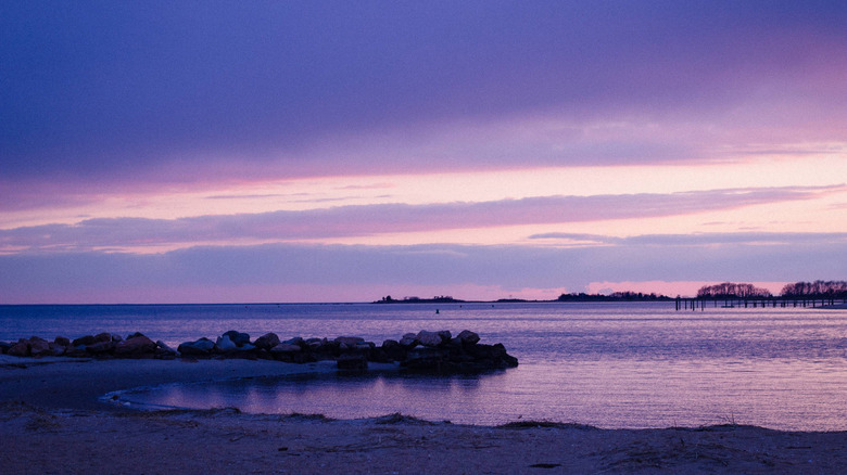 A purple beach and sky with a dark distant shoreline and low-lying violet stratus clouds before a peach and pink sky at Clinton Town Beach in Middlesex County, Connecticut