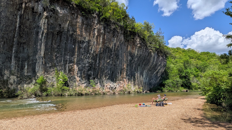 Two people sitting beside Sinking Creek with a towering concave bluff with foliage in Echo Bluff State Park