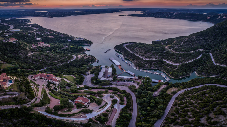 Aerial view of Lake Travis at sunset, with a marina and red-roofed white buildings on the close shore and treed lands dotted with homes around the lake