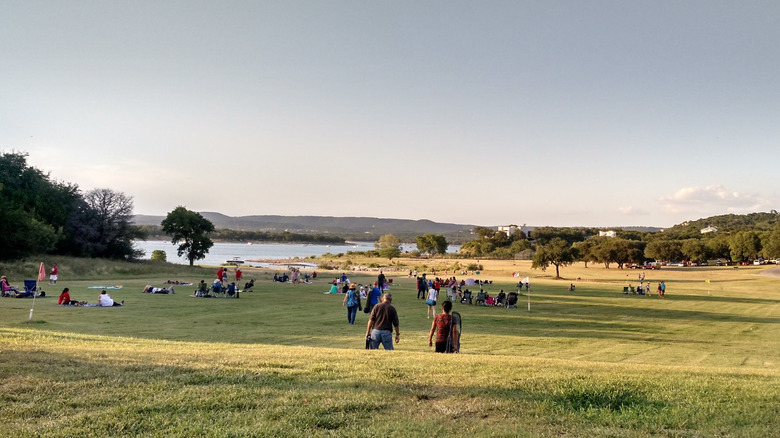 People walking and sitting in groups on a golf green, with a lake in the background surrounded by treed hills