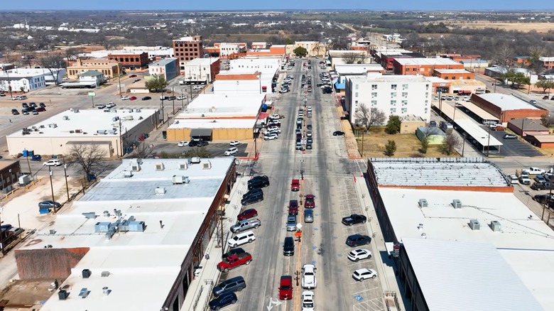 An aerial view of Commercial Avenue in downtown Coleman, Texas, showing a wide main street lined with historic storefronts, parked cars, and surrounding rural Texas landscape