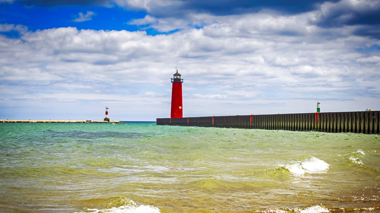 A small red and white striped lighthouse on a rocky jetty beside a red lighthouse at the end of a concrete boardwalk sitting in teal blue clear lake waters